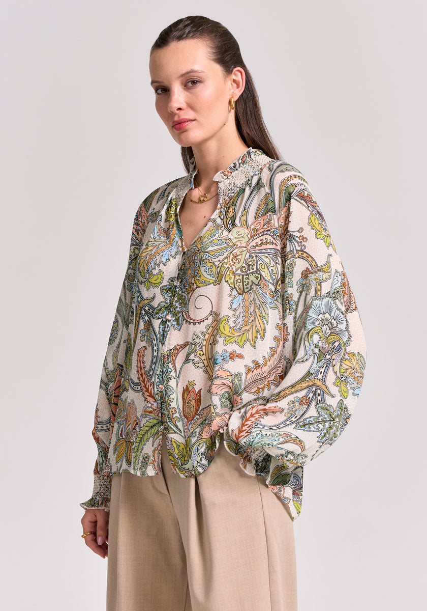 A woman stands against a plain background wearing a Princess Goes Hollywood Paisley-Print Bluse with Ballonärmel, ruffled neckline, and beige wide-leg trousers. Her straight brown hair is pulled back, her expression neutral, and she wears minimal jewelry.
