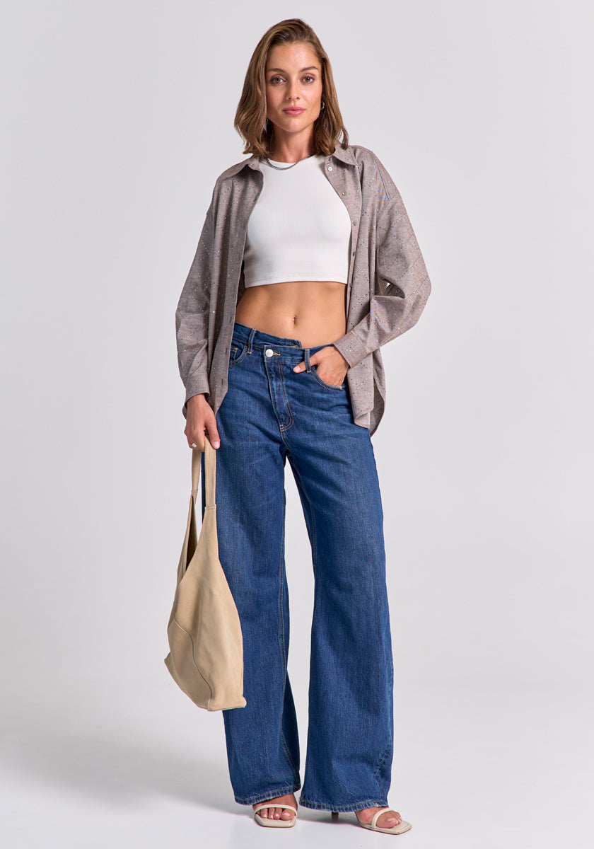 Eine Frau trägt ein weißes Baumwoll-Crop-Top, eine lockere Hemdbluse, blaue Wide-Leg-Jeans und Sandalen von Princess Goes Hollywood. In der linken Hand hält sie eine beige Tasche und blickt entspannt in die Kamera.