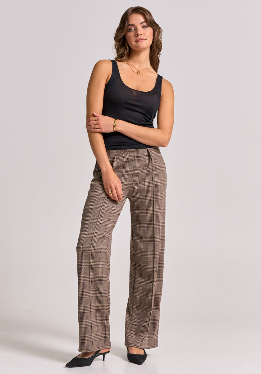 A woman stands confidently against a light gray background, wearing a black tank top, gold jewelry, and Princess Goes Hollywood’s white check-patterned trousers with an elastic waistband. She smiles gently, her wavy brown hair falling to her shoulders.