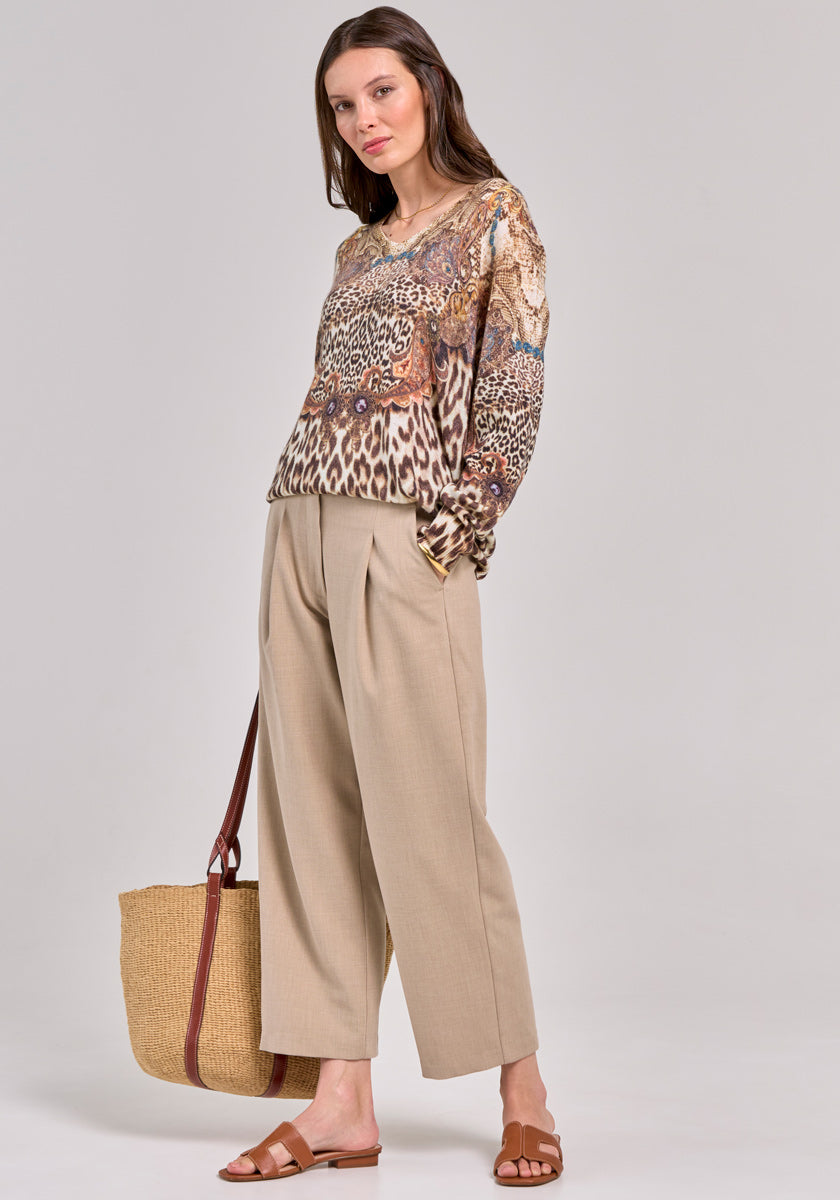 A woman wearing a Princess Goes Hollywood long-sleeve animal print blouse and beige wide-leg 7/8-length trousers stands against a plain background, styled with brown sandals and a large straw tote, looking at the camera with a relaxed posture.
