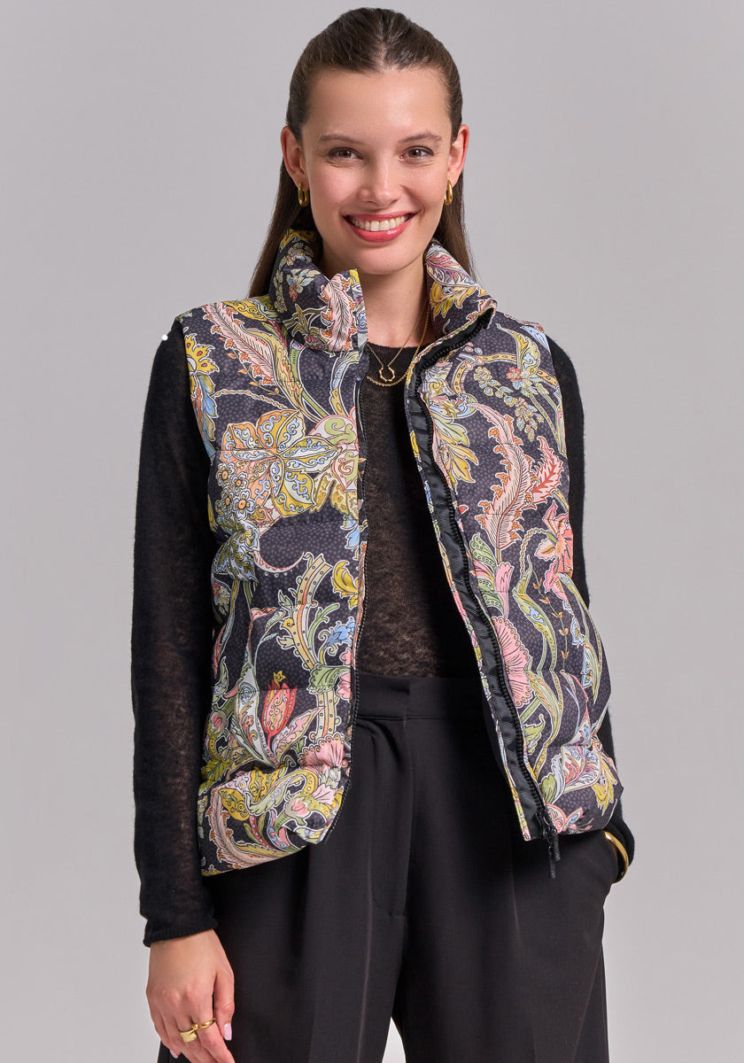 A woman with long brown hair, pulled back, smiles while wearing a Princess Goes Hollywood Paisley-Print Steppweste with two-way zipper over a black long-sleeve top and pants. She accessorizes with gold hoop earrings and a necklace. Light gray background.