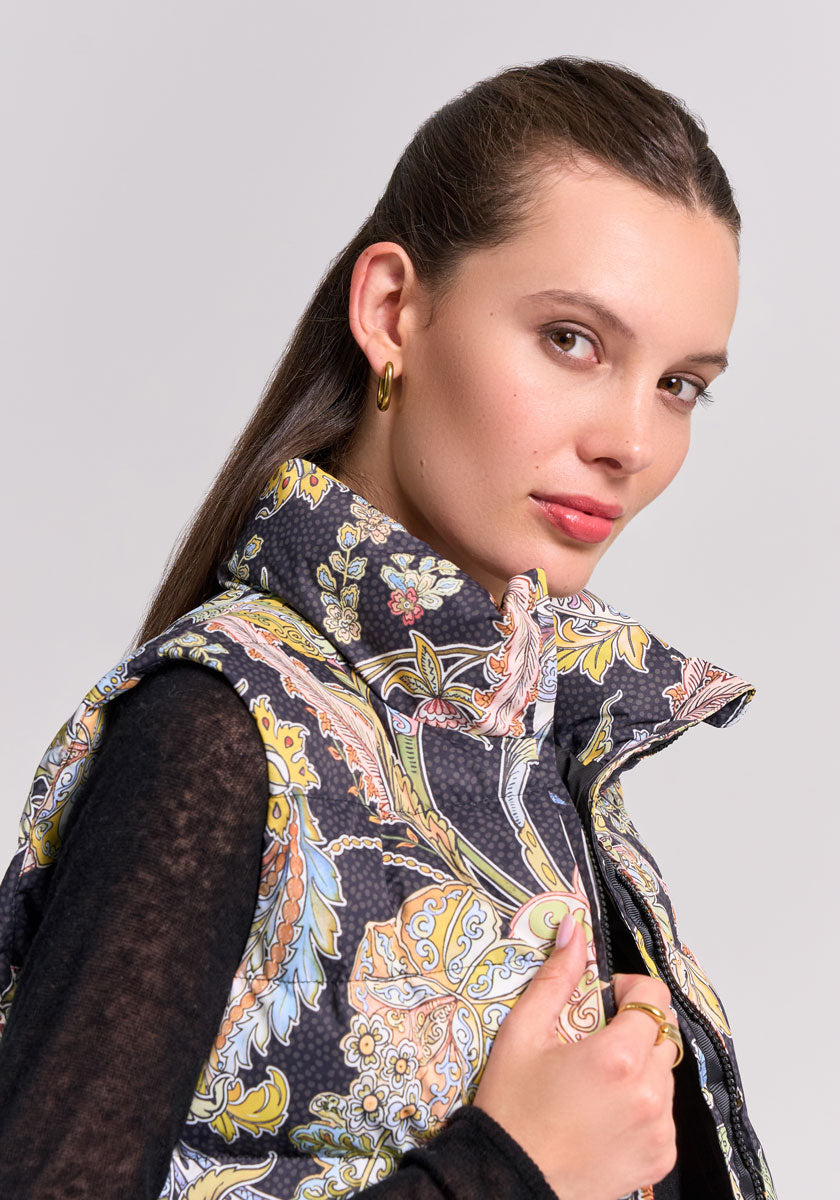 A woman with long brown hair wears gold hoop earrings and a Princess Goes Hollywood Steppweste with yellow, blue, and orange paisley print. She holds the collar, looking confidently at the camera against a light gray background.