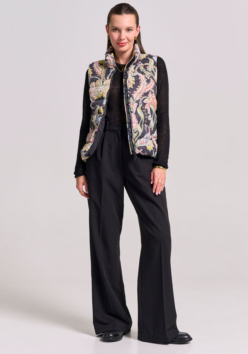 A woman stands facing forward, wearing a sheer black long-sleeve top, loose black trousers, black shoes, and a colorful paisley-print Steppweste by Princess Goes Hollywood. Her straight hair is pulled back and she poses against a plain background.