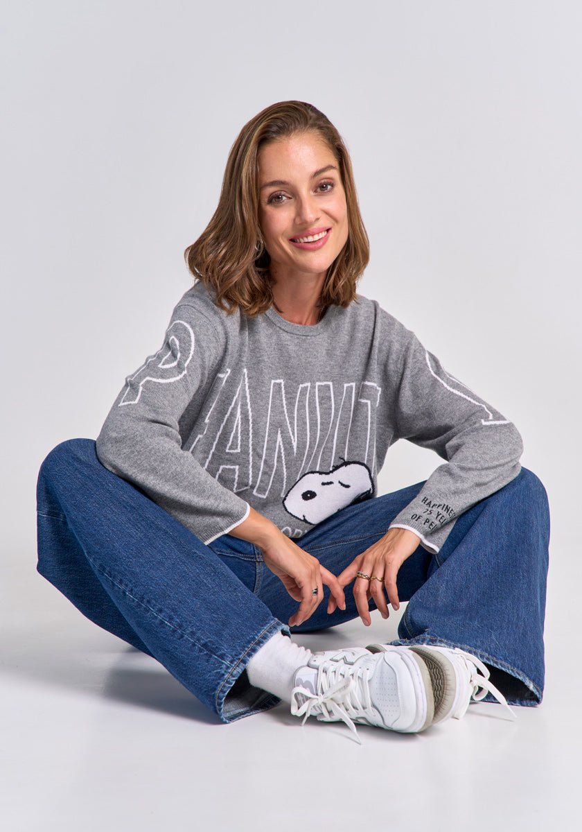 A woman with shoulder-length brown hair smiles while sitting on the floor, wearing a gray Peanuts Pullover mit Snoopy-Motiv by Princess Goes Hollywood, blue jeans, white socks, and sneakers. She sits cross-legged with her left arm resting on her knee.