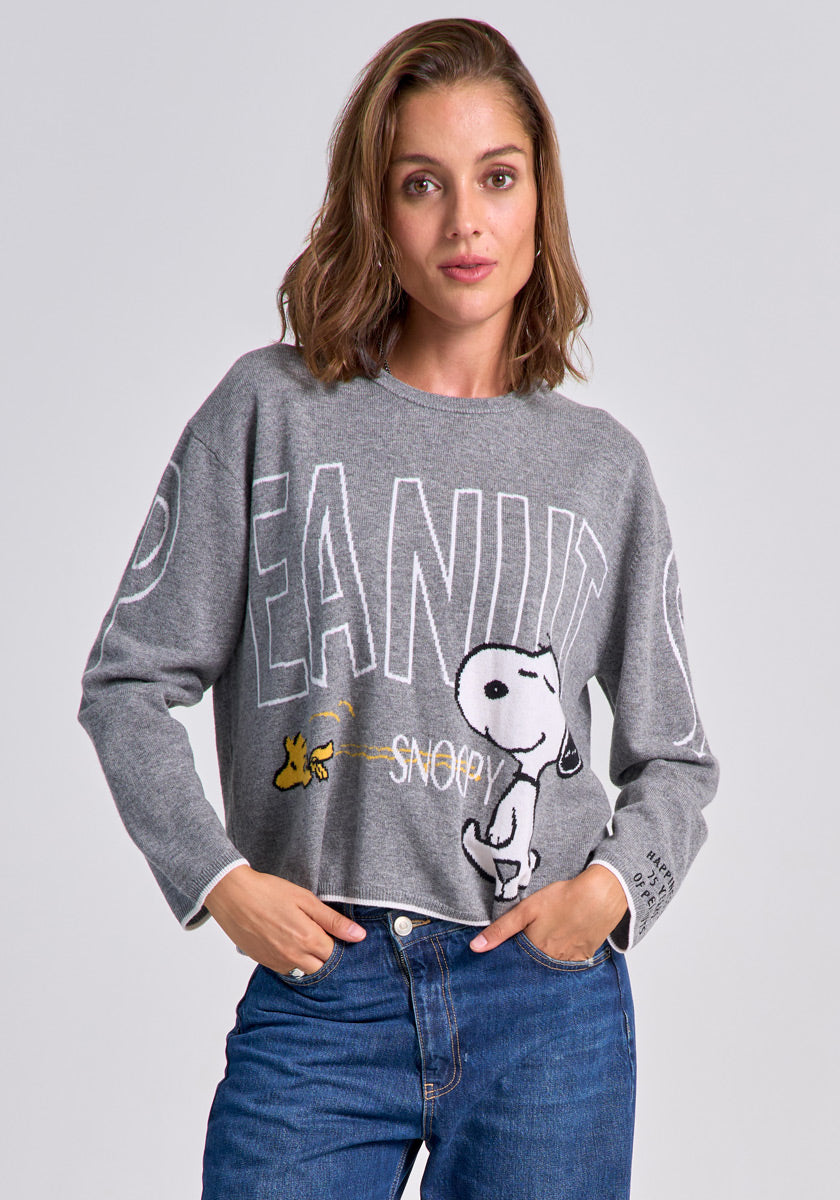 A woman with shoulder-length brown hair models a grey Princess Goes Hollywood Peanuts Pullover featuring Snoopy, Woodstock, and bold “PEANUT” text. She wears blue jeans and stands with hands in her pockets against a plain grey background.