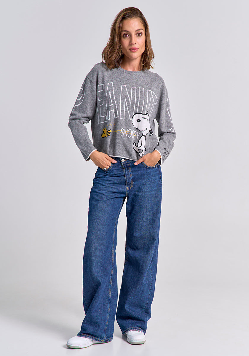 A woman with shoulder-length brown hair wears a grey Princess Goes Hollywood Peanuts Snoopy Pullover with Snoopy and Woodstock graphics, paired with wide-leg blue jeans and white sneakers, standing relaxed against a light grey background.