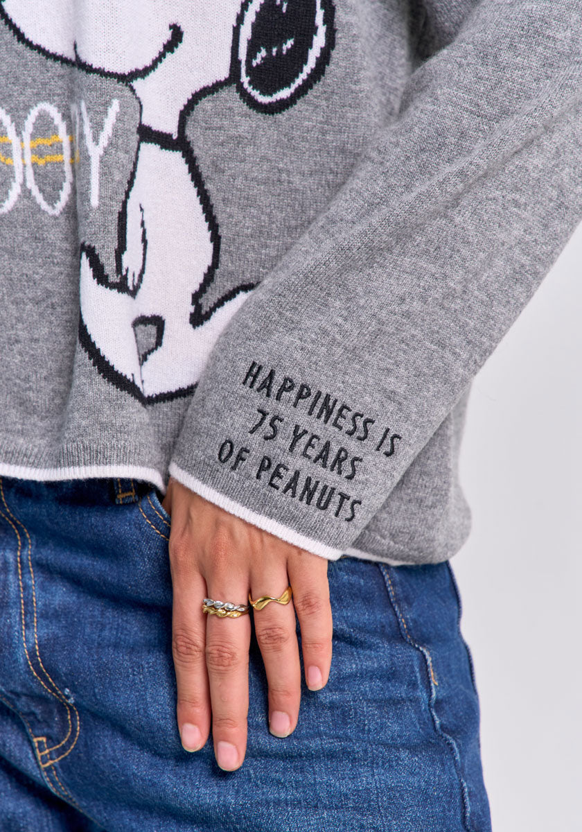 A person wears blue jeans and a grey Princess Goes Hollywood Peanuts Snoopy pullover showing part of Snoopy’s face. Their left hand, with two rings, rests on their thigh. The sweater cuff reads, “Happiness is 75 years of Peanuts.”.