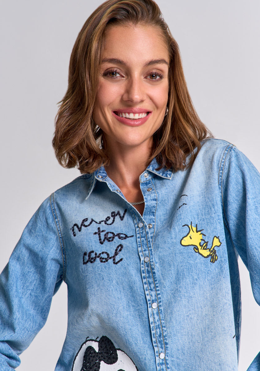 A smiling woman with light brown hair wears a Princess Goes Hollywood light blue Baumwolle Jeanshemd featuring a Snoopy and Woodstock print, with "never too cool" embroidered in black on the chest. The background is plain, light gray.