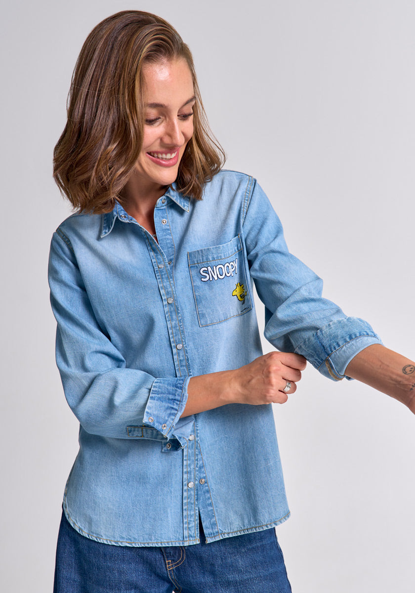 A woman with shoulder-length brown hair smiles as she rolls up the sleeve of her light blue Princess Goes Hollywood cotton Jeanshemd, featuring a chest pocket with Snoopy embroidery and a small Woodstock image, against a plain background.