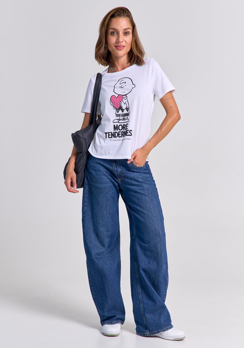 A woman wearing a white Princess Goes Hollywood Charlie Brown cotton T-shirt with a cartoon and "More Tenderness" text, blue wide-leg jeans, and white sneakers stands against a plain background, carrying a dark shoulder bag and smiling slightly.