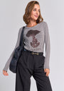 A woman with wavy, shoulder-length hair wears a Princess Goes Hollywood striped long-sleeve shirt with Snoopy motif and sequin details, tucked into belted high-waisted black trousers. She carries a black padded bag on her shoulder and smiles, looking to the side.