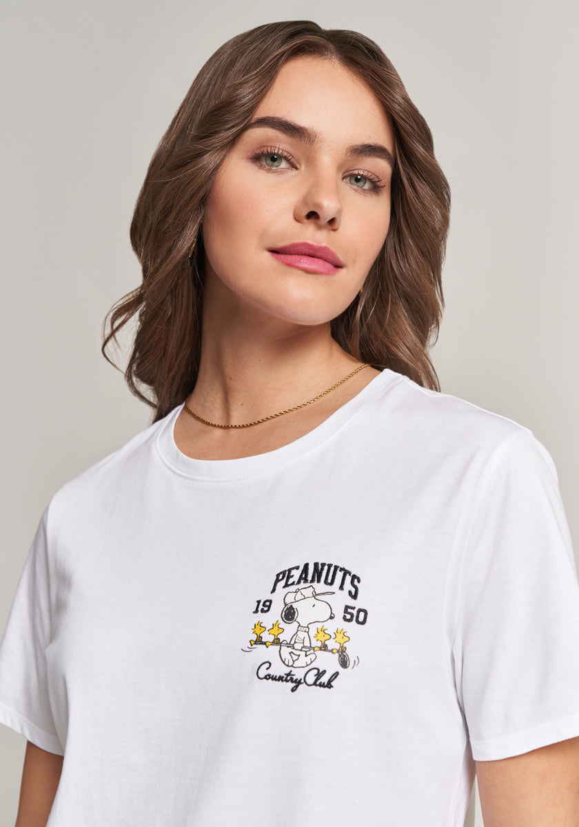 A woman with wavy brown hair wears a white Frogbox by Princess Baumwolle T-shirt with a small Snoopy and Woodstock print, reading Peanuts Country Club. She stands against a plain background, accessorized with a gold chain necklace.