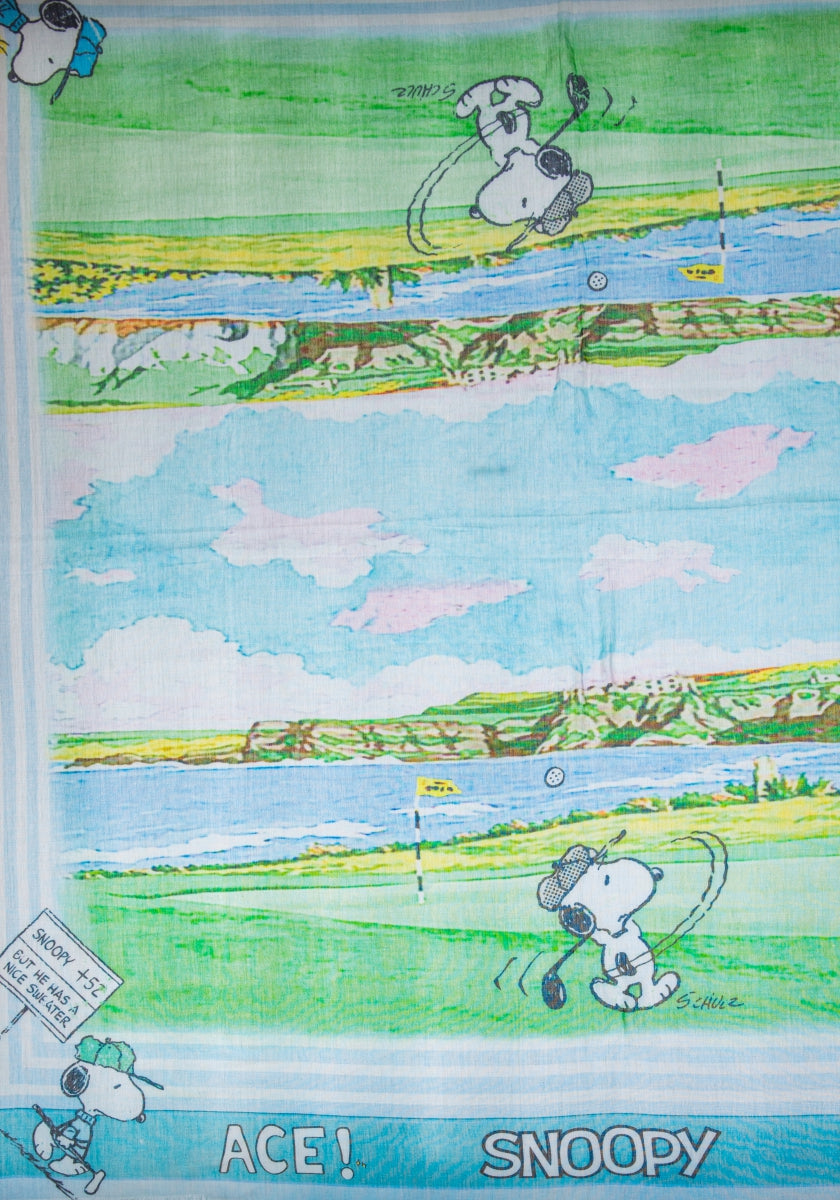 A colorful Frogbox by Princess Snoopy-print scarf shows Snoopy golfing on green grass near a lake and trees under a blue sky. Made from a cotton-modal mix, it features “ACE! SNOOPY” and a sign with Snoopy 72, Handicap 0.