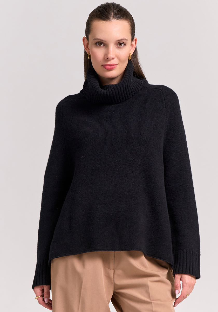 A woman with long brown hair wears a Frogbox by Princess schwarzer Merinowolle Pullover mit Rollkragen und Raglanärmel, paired with tan trousers. She stands against a plain light background, facing the camera with a neutral expression.