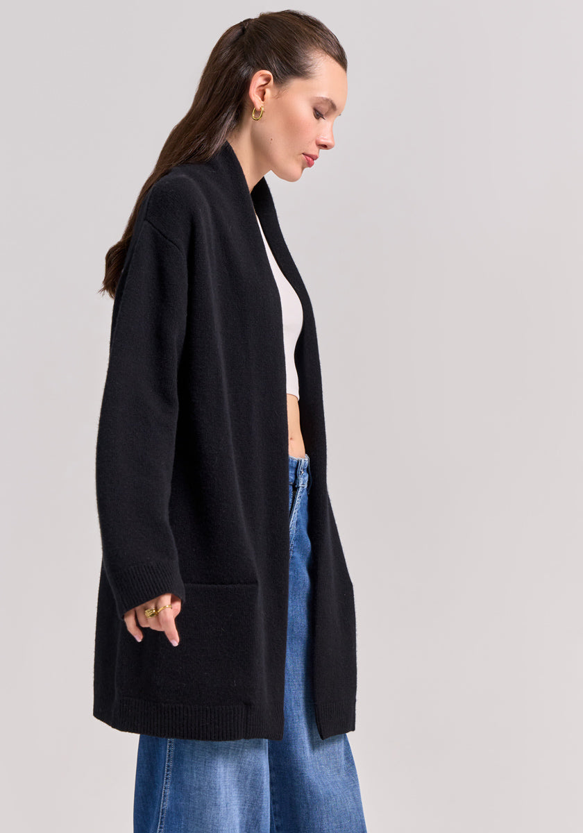 A woman with long brown hair, styled half-up, wears a black Frogbox by Princess Strickjacke with Schalkragen over a white crop top and blue wide-leg jeans. She stands in profile against a plain light background, looking down with one hand at her side.