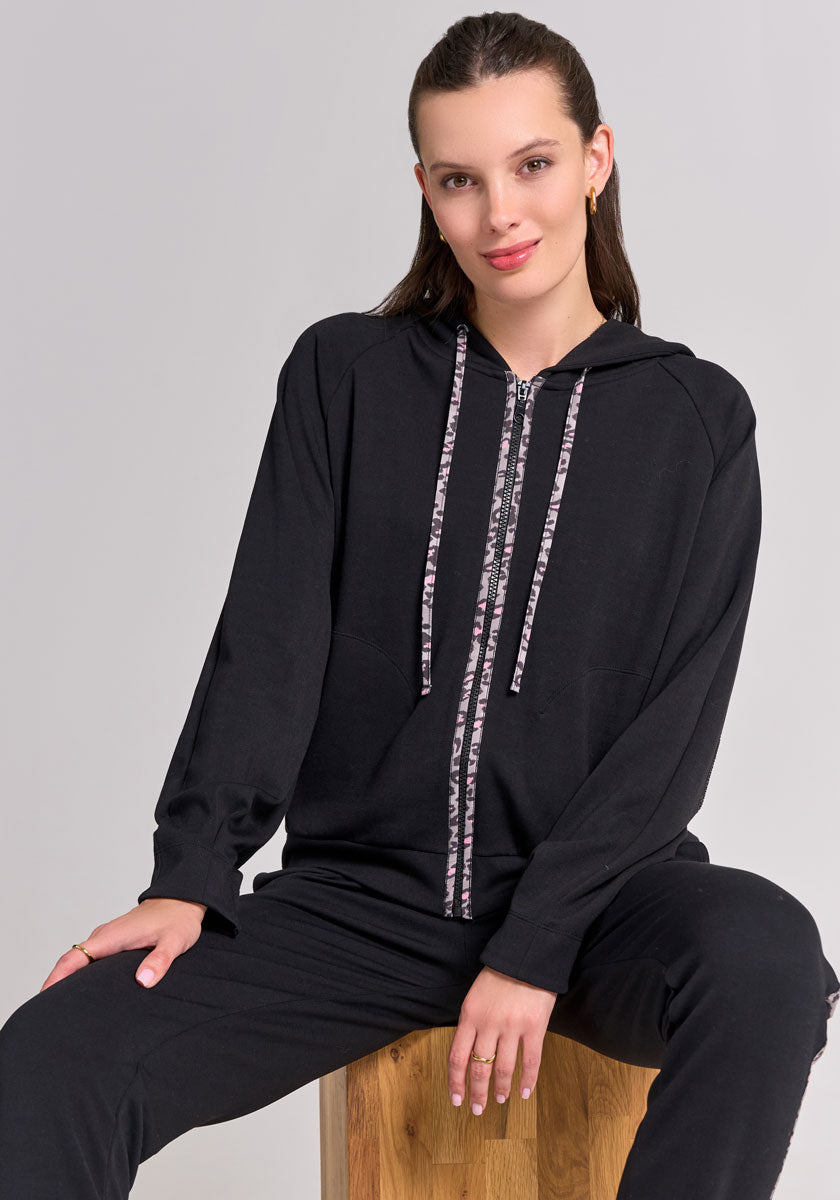 A person with straight brown hair, wearing a Frogbox by Princess black hooded zip-up jacket with patterned drawstrings and matching black Sweathose pants, sits on a wooden stool against a light gray background, looking at the camera and slightly smiling.