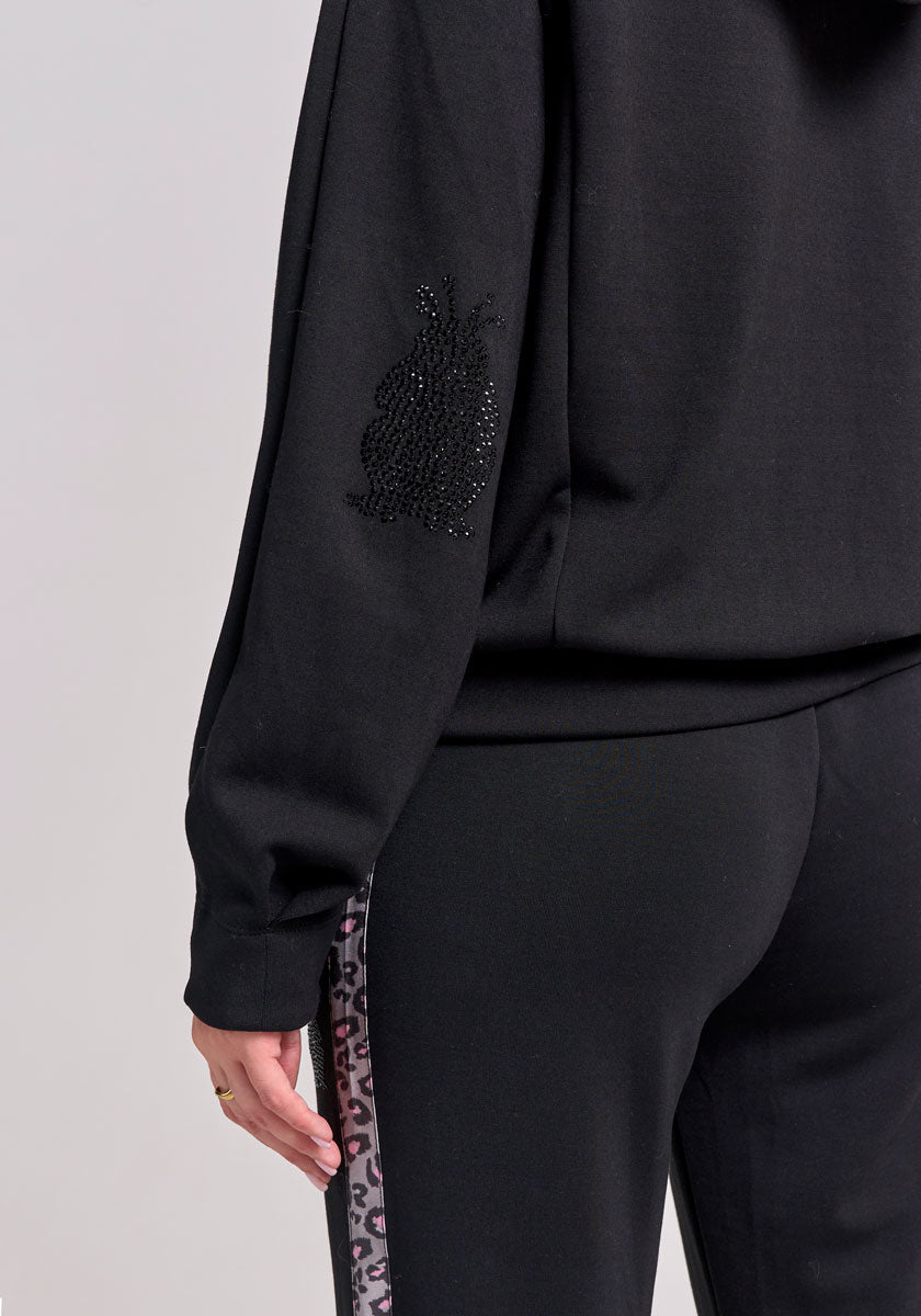 A person in a black Frogbox by Princess outfit is seen from the back and side, wearing sweathose with a pink and grey leodetail animal print stripe, a sleeve adorned with a black beaded owl patch, and a gold ring on their hand.