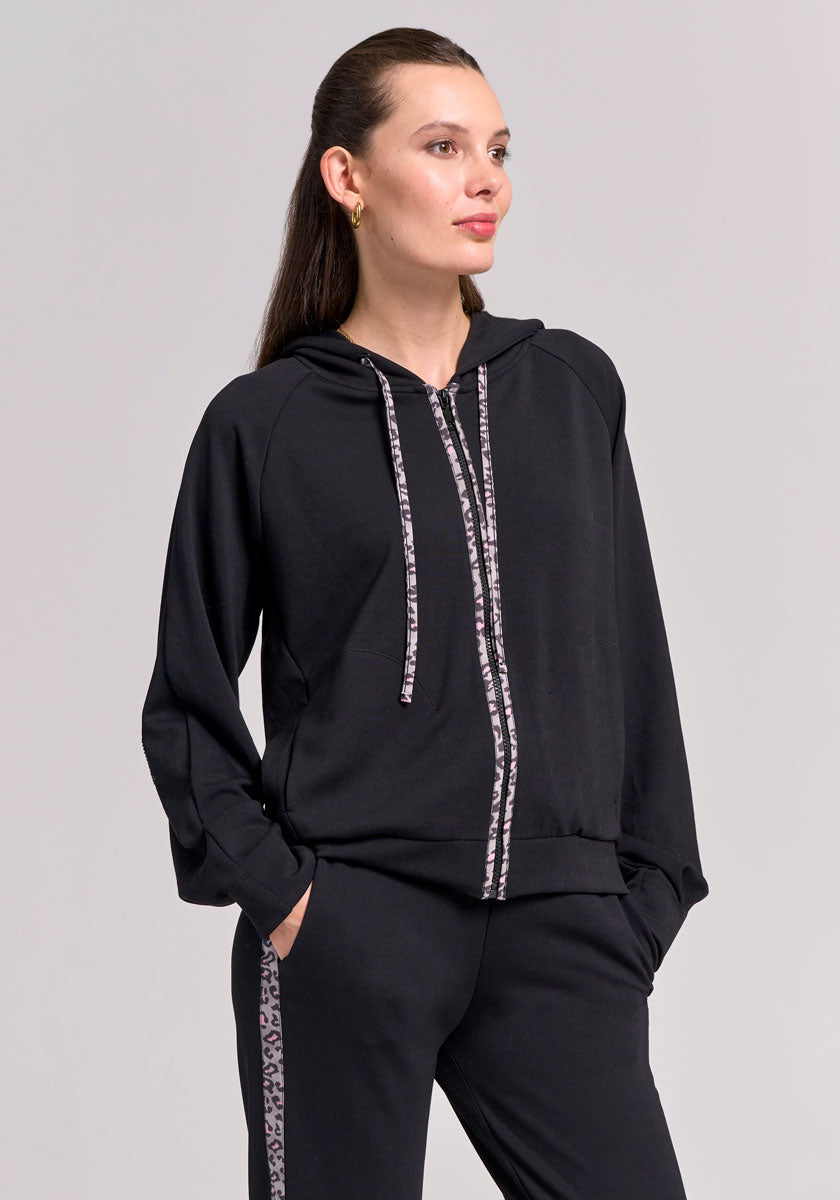 A woman with long brown hair in a ponytail and gold hoop earrings poses against a light gray background, wearing a Frogbox by Princess black zip-up hoodie with patterned drawstrings and matching Sweathose with Leodetail side stripes.
