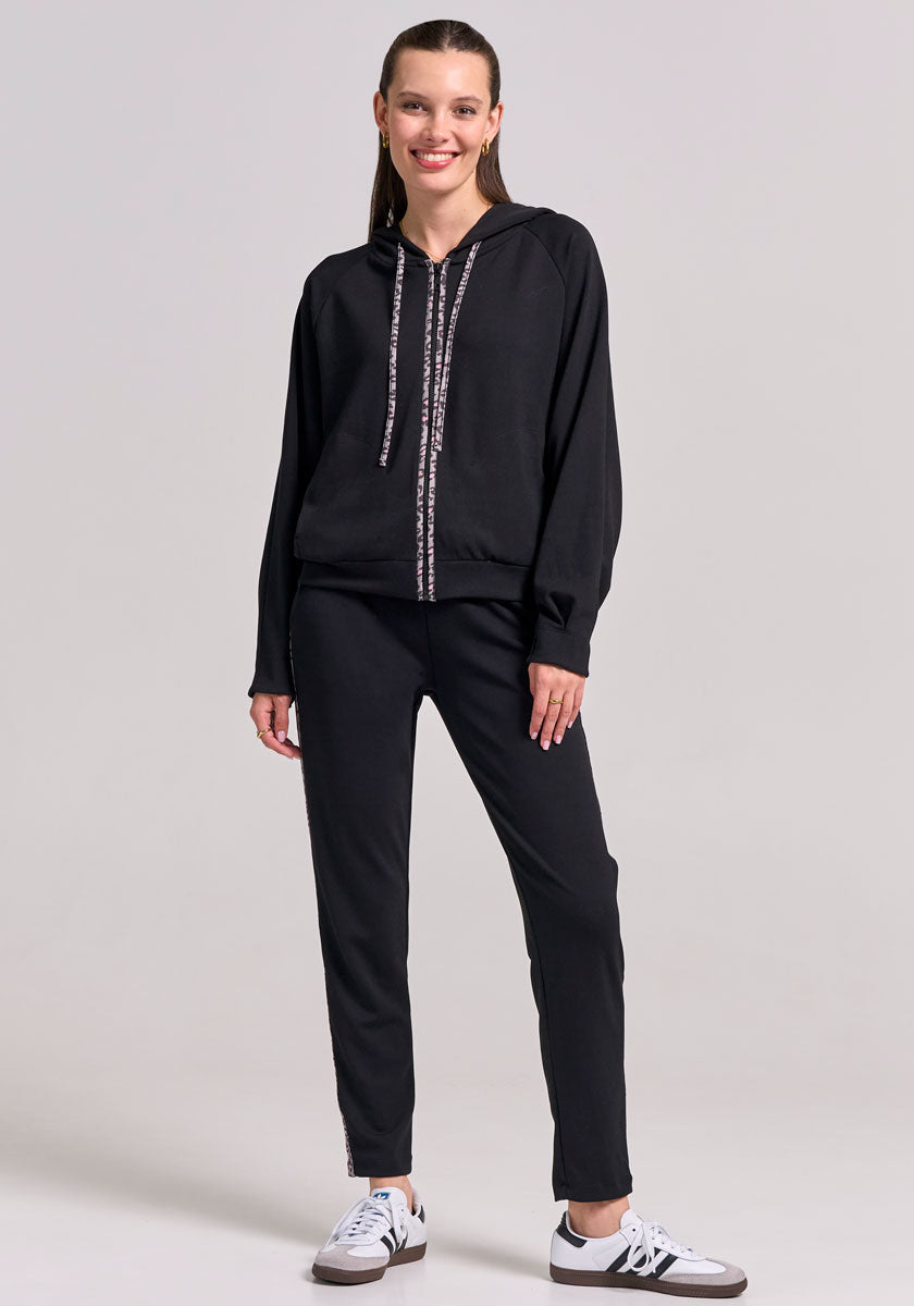 A smiling woman with straight, light brown hair stands casually in Frogbox by Princess: a matching black hoodie and sweathose with drawstrings, plus white sneakers with black accents, against a plain background.