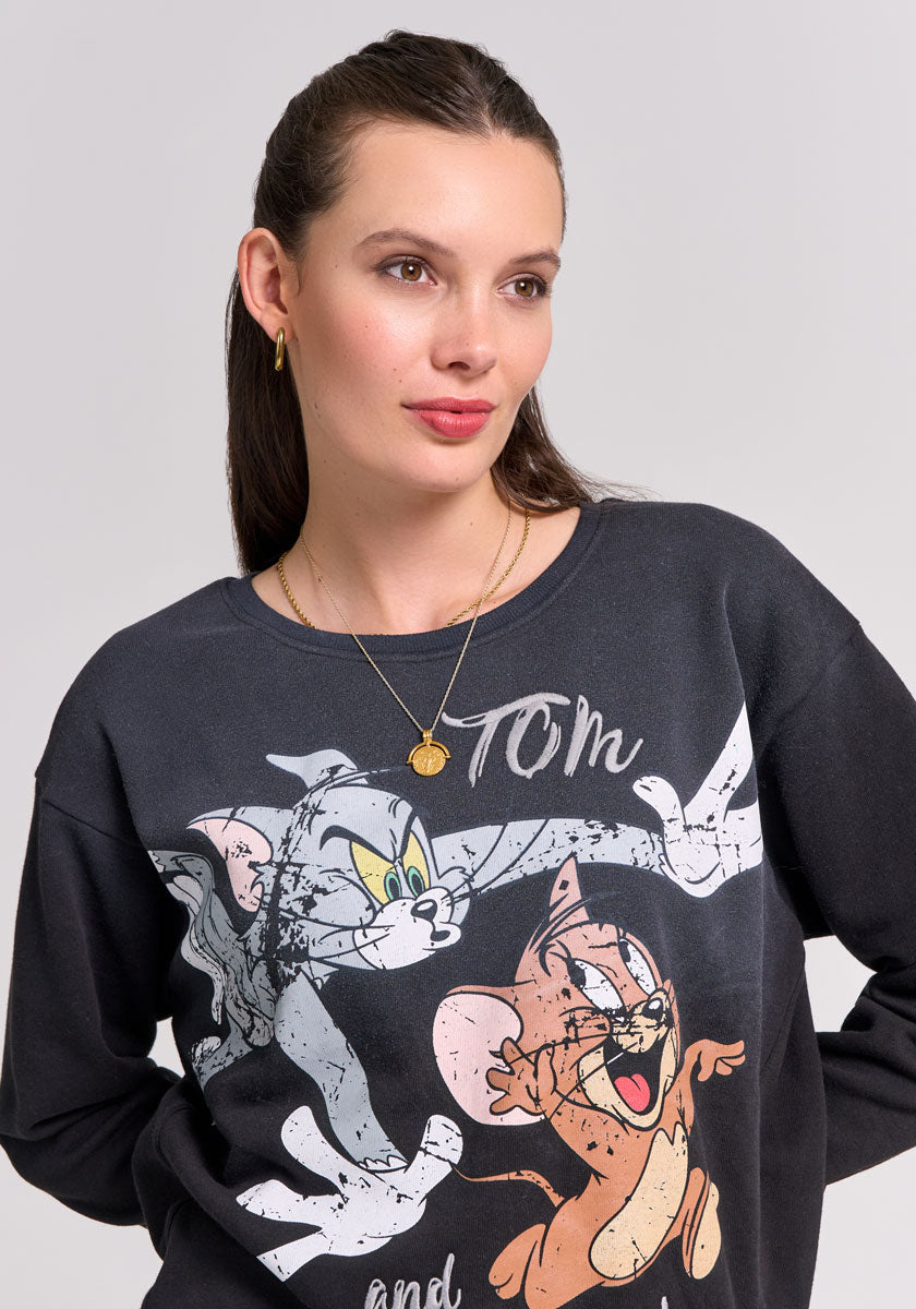 A woman with fair skin and brown hair tied back wears gold jewelry and a black Frogbox by Princess Rundhals Sweatshirt. She gazes slightly to the side, her neutral expression highlighting the playful sweatshirt against a plain background.