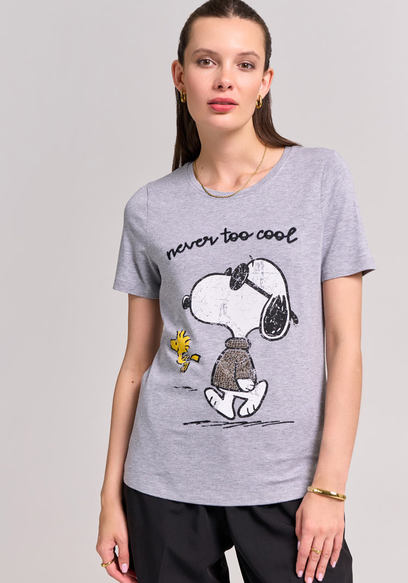 A woman with straight brown hair wears a Frogbox by Princess Snoopy T-shirt with Woodstock and never too cool text, styled with gold jewelry and black pants against a plain light background.