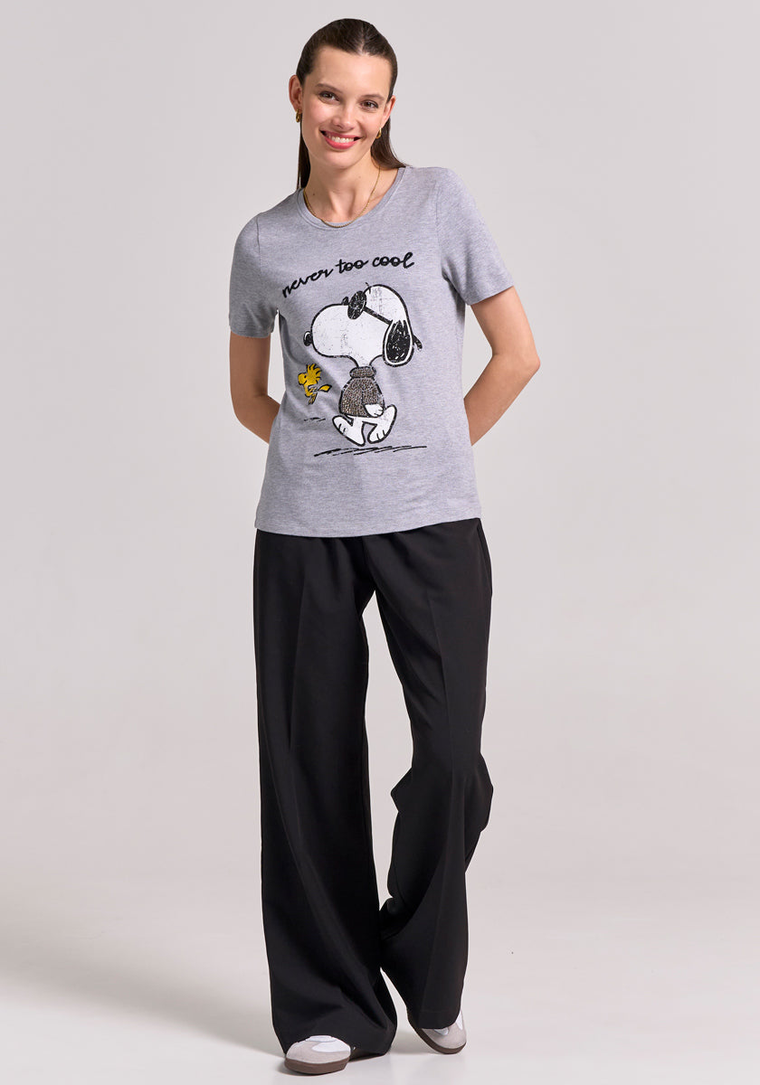 A smiling woman stands against a plain light background, wearing a Frogbox by Princess grey Snoopy T-shirt with never too cool text and Rundhalsausschnitt, black wide-leg pants, and white sneakers. Her brown hair is pulled back.