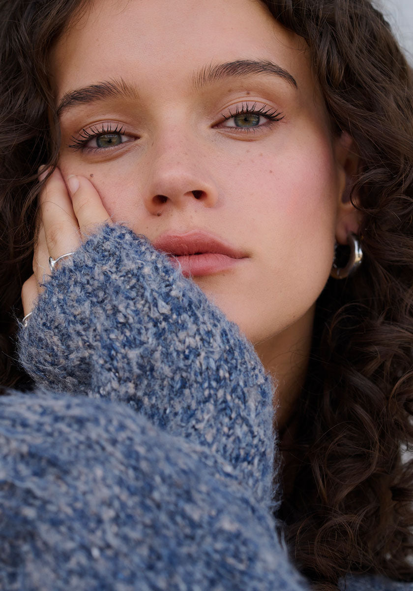 Eine junge Frau mit lockigen braunen Haaren und grünen Augen blickt sanft in die Kamera, ihre Hand ruht auf ihrem Gesicht und wird teilweise von einem strukturierten blauen Strickpullover aus Seide mit Raglanärmeln von Princess Goes Hollywood verdeckt. Sie trägt silberne Ohrringe und minimales Make-up.