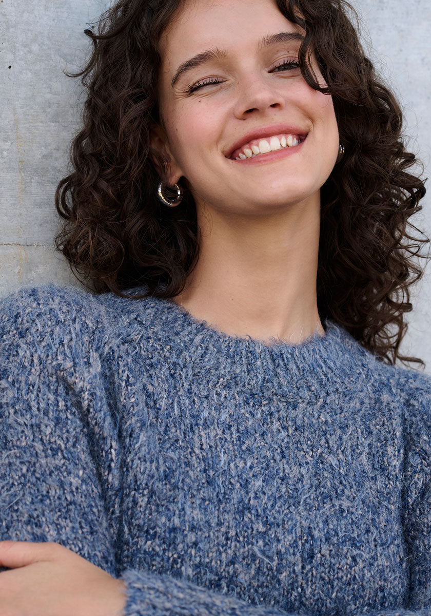 Eine junge Frau mit lockigem braunem Haar lächelt breit, trägt silberne Ohrringe und einen blauen Strickpullover mit Rundhalsausschnitt von Princess Goes Hollywood. Der weiche, neutrale Hintergrund unterstreicht ihren strahlenden Ausdruck.