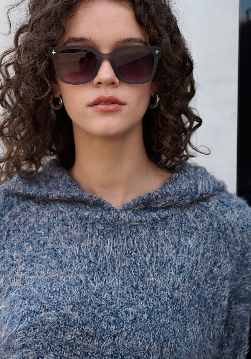 Eine Frau mit lockigem braunem Haar trägt eine dunkle Sonnenbrille, silberne Ohrringe und einen strukturierten blauen Strickhoodie mit Kapuze und Raglanärmel von Princess Goes Hollywood. Sie steht im Freien und schaut bei natürlichem Licht selbstbewusst in die Kamera.