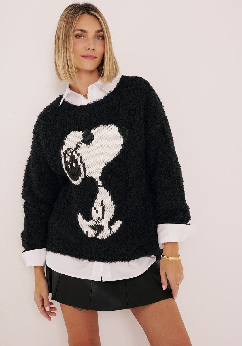 Eine Frau mit blondem Bob modelliert einen schwarzen Princess Goes Hollywood Kuschelpullover mit einem großen handgestricktes Snoopy-Motiv, gestylt über einem weißen Hemd mit Kragen und einem kurzen schwarzen Rock, der ruhig vor einer schlichten hellen Wand steht.