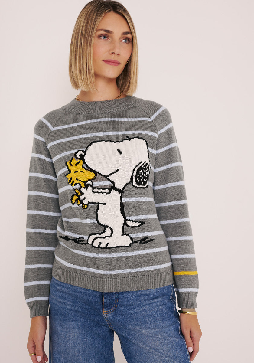 Eine Frau mit glattem blondem Haar trägt einen gestreiften Baumwollpullover aus der Serie Princess Goes Hollywood mit einer großen Snoopy-Woodstock-Grafik, dazu blaue Jeans. Sie steht vor einem schlichten, hellen Hintergrund und sieht entspannt und neutral aus.