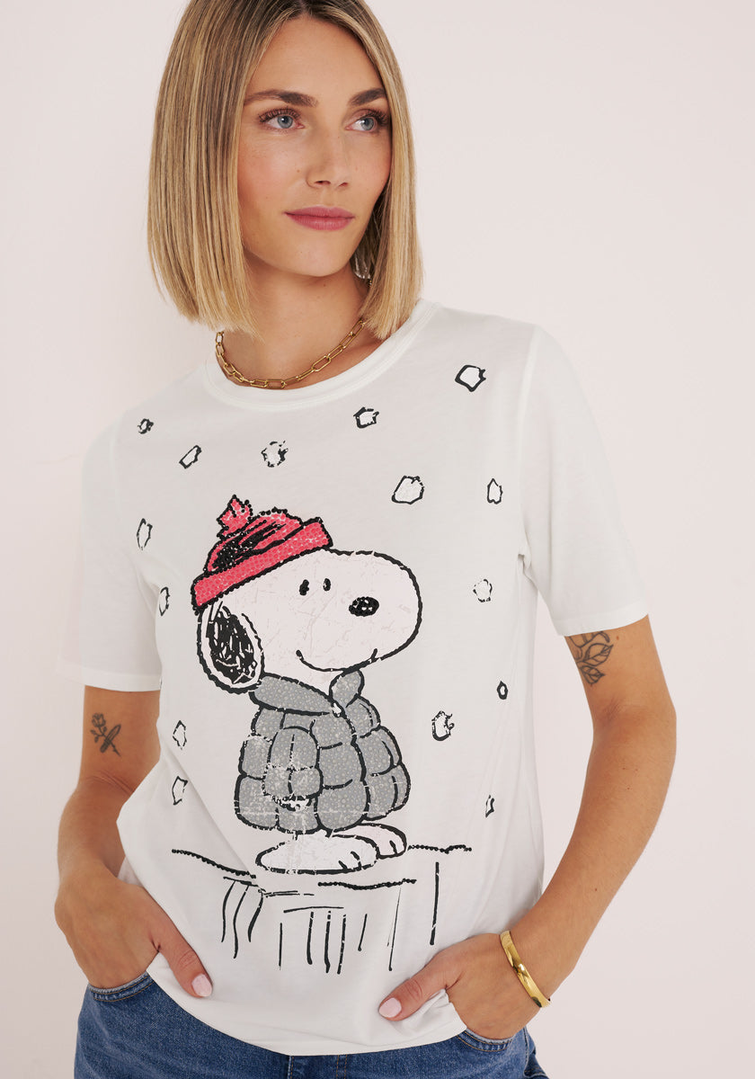 Eine Frau mit blondem Haarschopf trägt ein Princess Goes Hollywood Snoopy T-Shirt aus Baumwolle, das Snoopy mit rotem Hut und bauschiger Jacke mit Schnee zeigt. Sie steht vor einem schlichten, hellen Hintergrund mit einer Hand in der Tasche und einem entspannten Gesichtsausdruck.