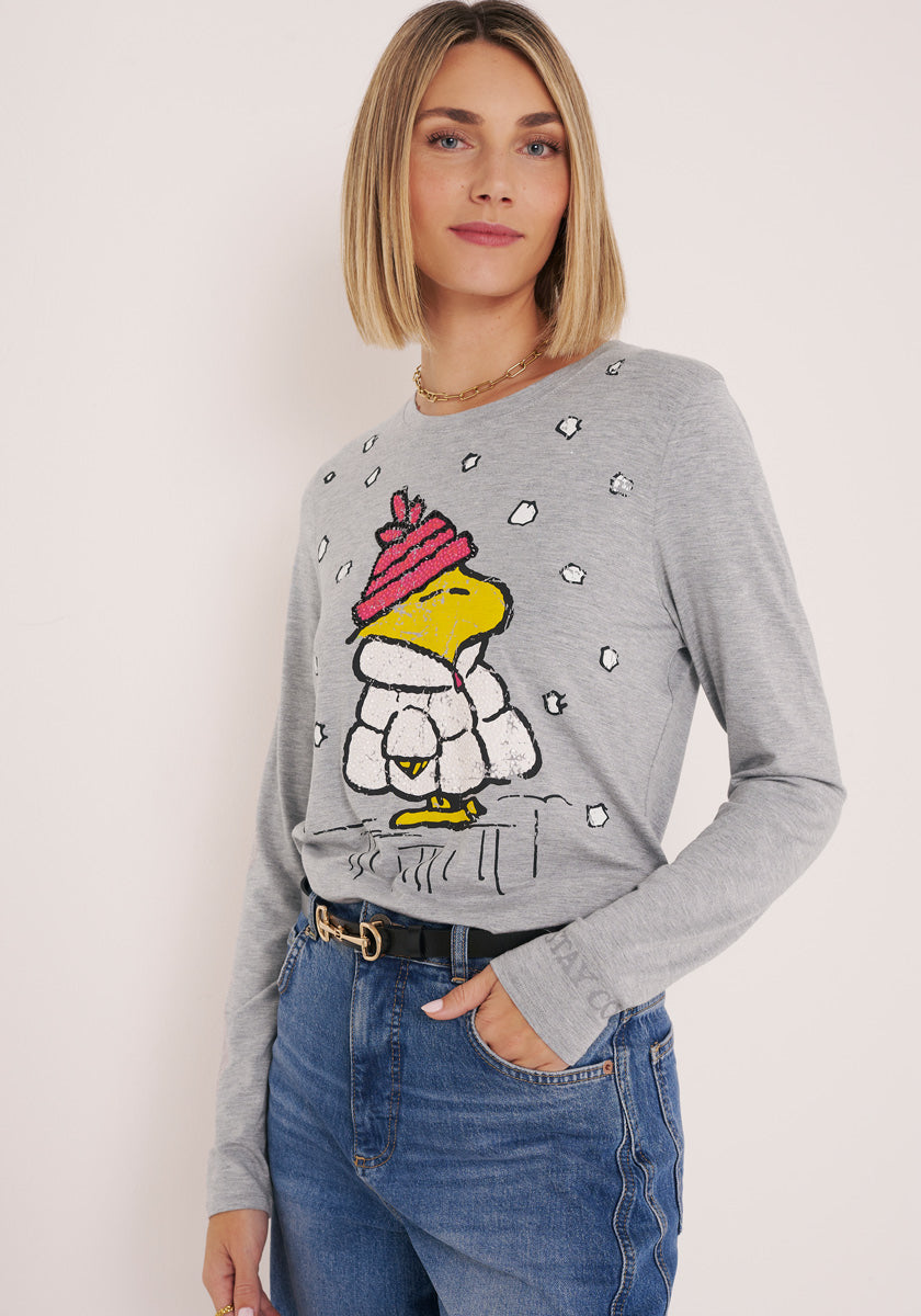 Eine Frau mit blondem Haarschopf trägt ein hellgraues Princess Goes Hollywood Langarmshirt mit einem Woodstock-Cartoon-Vogel mit rosa Hut und weißem Mantel inmitten von Schneeflocken, gepaart mit blauen Jeans und einem schwarzen Gürtel, vor einem schlichten Hintergrund.