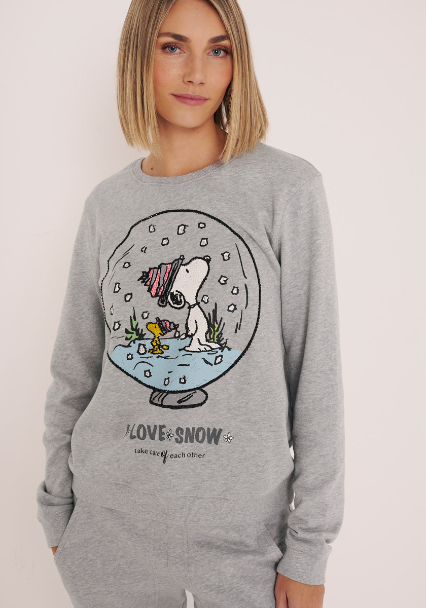 Eine Frau mit glattem blondem Haar trägt ein hellgraues Princess Goes Hollywood Baumwolle Damen Sweatshirt mit Snoopy und Woodstock in einer Schneekugel, funkelnder Strassverzierung und dem Schriftzug "LOVE SNOW, take care of each other".