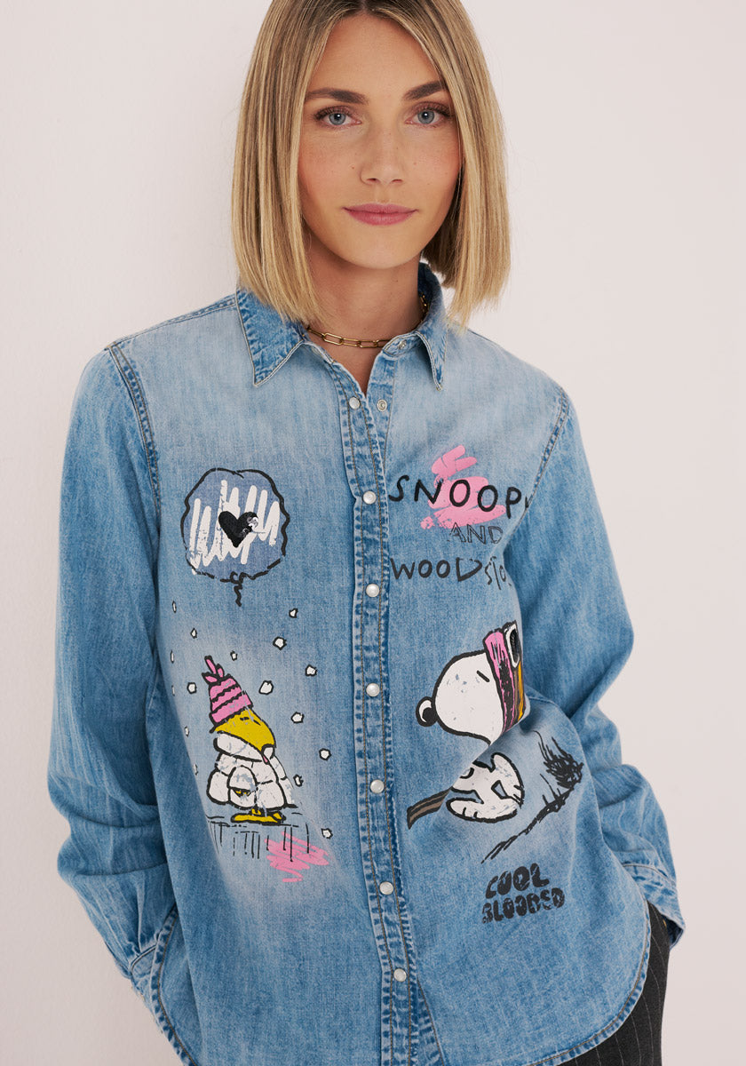 Eine Frau mit glattem blondem Haar trägt ein Princess Goes Hollywood-Jeanshemd aus Baumwolle mit bunten Snoopy- und Woodstock-Aufdrucken. Sie steht vor einem schlichten, hellen Hintergrund und blickt mit einem leichten Lächeln selbstbewusst in die Kamera.