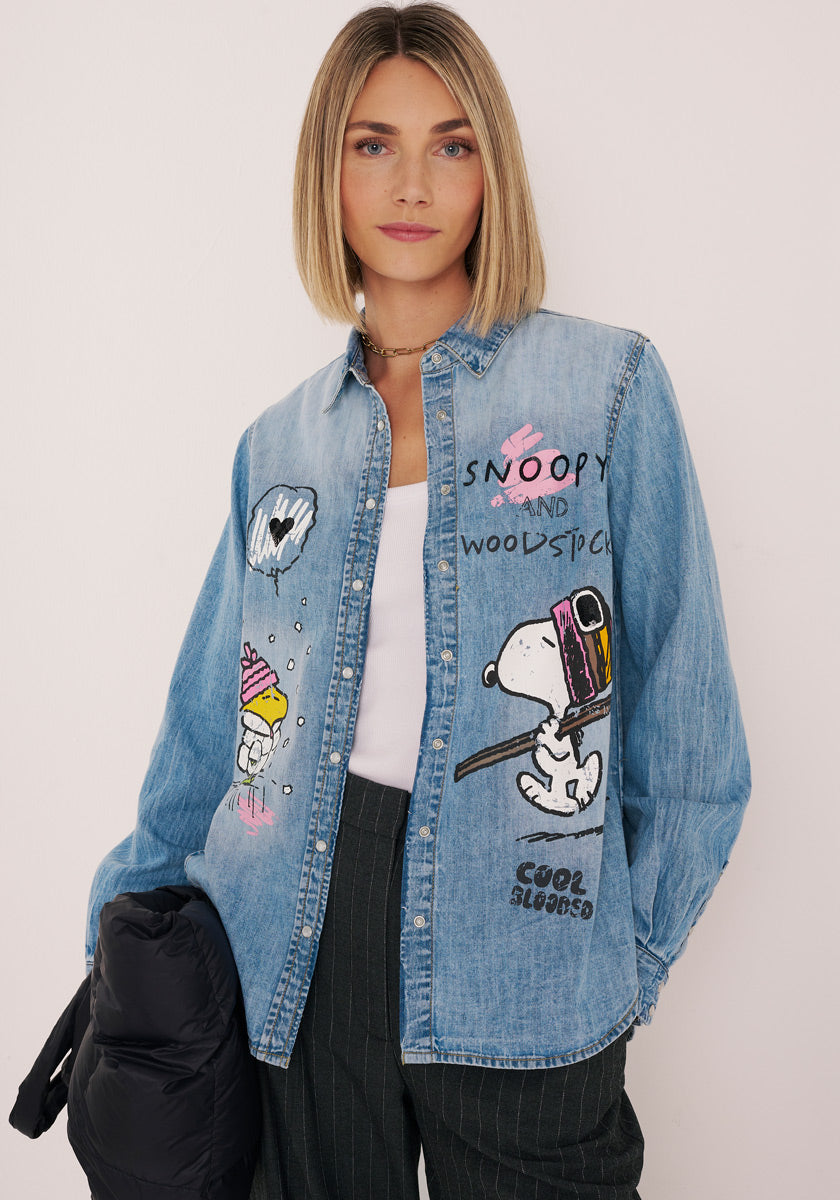 Eine Frau mit glattem blondem Haar trägt ein helles Baumwoll-Jeanshemd mit Snoopy-Aufdruck von Princess Goes Hollywood, auf dem verspielte Peanuts-Figuren abgebildet sind. Dazu trägt sie ein weißes Oberteil, eine dunkle Hose mit Nadelstreifen und eine schwarze Jacke.