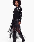 Eine Frau steht in einem schwarzen Pullover mit weißen Sternen, dazu trägt sie einen Minirock aus veganem Leder mit langen Fransen und kniehohe schwarze Stiefel. Sie hat eine natürliche Afro-Frisur und trägt Creolen, während sie selbstbewusst in die Kamera blickt. Der Hintergrund ist weiß.
