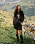 Eine Frau steht im Freien auf grasbewachsenen Hügeln, im Hintergrund sind Berge zu sehen. Sie trägt eine braune Jacke, einen Pullover aus Alpaka-Wolle mit Snoopy-Intarsien im Animal-Print, einen schwarzen Lederrock und schwarze Stiefel. Sie blickt leicht nach oben, ihr langes Haar fällt ihr offen ins Gesicht, die Hände hält sie an den Seiten. Neben ihr steht ein Holzzaun.