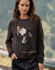 Eine Frau mit welligem braunem Haar trägt einen dunklen Snoopy-Intarsia-Pullover, auf dem Snoopy und Woodstock abgebildet sind, zusammen mit dem Schriftzug „Peanuts“ und „Have a wonderful day“. Sie steht draußen neben einem Holzgeländer, im Hintergrund sind verschwommene grüne Berge und Bäume zu sehen.