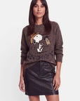 Eine Frau in einem braunen Pullover aus Alpaka-Wolle mit Snoopy-Intarsien und dezenten Strassverzierungen steht mit dem Blick nach vorne. Ihre Hände ruhen an den Seiten. Sie hat langes, glattes, dunkles Haar und einen neutralen Gesichtsausdruck. Der Hintergrund ist schlicht weiß.