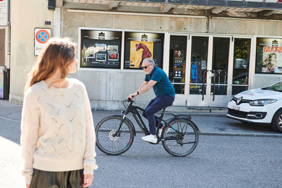 A man in sunglasses rides a black bicycle on a street, glancing toward a woman in a cream sweater standing in the foreground. Behind them is a building with posters and a parked white car. The scene is in daylight on a city street.