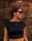 Eine Frau mit dunklem Haar in einem Dutt, geschmückt mit einer roten Blume, steht vor einer rustikalen Steinmauer. Sie trägt eine Okkia Oversized Sonnenbrille mit UV 400-Schutz, ein schwarzes Crop-Top, goldene Ohrringe, mehrlagige Ketten und ein Bauchnabelpiercing.