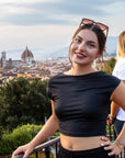 Eine lächelnde junge Frau posiert an einem Geländer in Florenz, Italien, und trägt eine übergroße Okkia Sonnenbrille, ein schwarzes Crop-Top und Reifohrringe. Die ikonische Kathedrale und das Stadtbild erscheinen hinter ihr unter einem bewölkten Himmel.