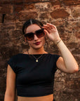 Eine Frau mit dunklem Haar in einem tiefen Dutt trägt eine Okkia Oversized Sonnenbrille mit UV 400-Schutz, ein schwarzes Crop-Top, goldene Halsketten und ein Armband. Sie steht selbstbewusst vor einer rustikalen Steinmauer, hält ihre Sonnenbrille und schaut in die Kamera.