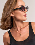 A woman with chin-length brown hair wears black Princess Goes Hollywood sunglasses, a black sleeveless top, and layered gold necklaces—a Schlangenkette and one with a Kreuzanhänger—looking to her right against a light background.