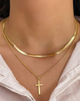 A close-up of a person’s neck wearing two gold Princess Goes Hollywood necklaces—a Schlangenkette choker and a thin Kette mit Kreuzanhänger—paired with a white collared shirt.