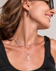 A woman with brown hair tucked behind her ear smiles, wearing black glasses, a black tank top, silver hoop earrings, and a dreischichtige Halskette in Silberoptik mit Kreuzanhänger von Princess Goes Hollywood.