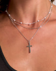 A close-up of a tan, smooth neck wearing a Princess Goes Hollywood dreischichtige Silberoptik-Halskette: one with pearls, one plain chain, and one with a minimalist Kreuzanhänger, styled with a black tank top.