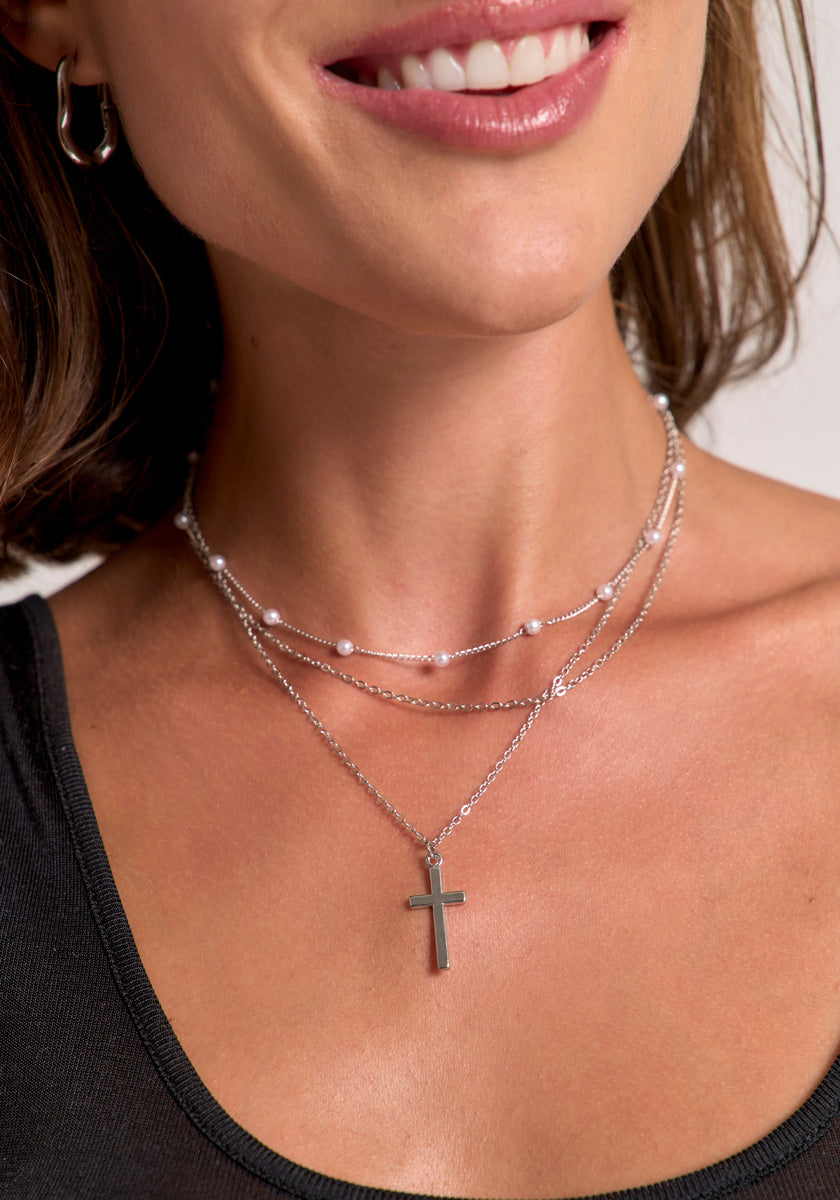 A woman with short brown hair smiles, revealing her teeth. She wears small hoop earrings and a silvery dreischichtige Halskette by Princess Goes Hollywood: one strand with a Kreuzanhänger, another with delicate white pearls.