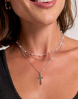 A woman with short brown hair smiles, revealing her teeth. She wears small hoop earrings and a silvery dreischichtige Halskette by Princess Goes Hollywood: one strand with a Kreuzanhänger, another with delicate white pearls.